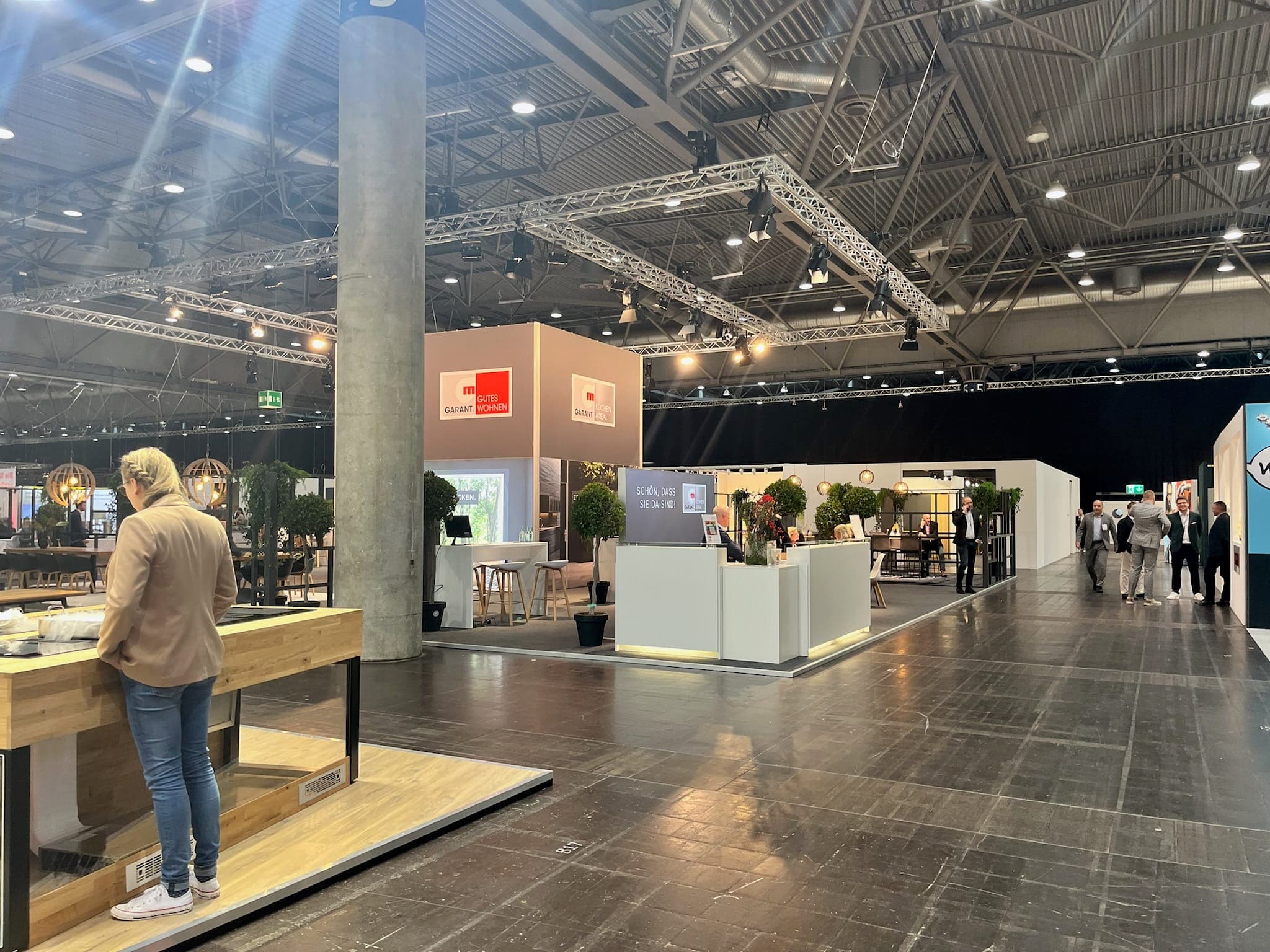 Innenaufnahme einer Messehalle mit modernen Ausstellungsständen zum Thema Wohnen und Einrichtung, darunter ein Stand, Besucher in Gesprächen sowie dekorative Pflanzen und stilvolle Möbelstücke.