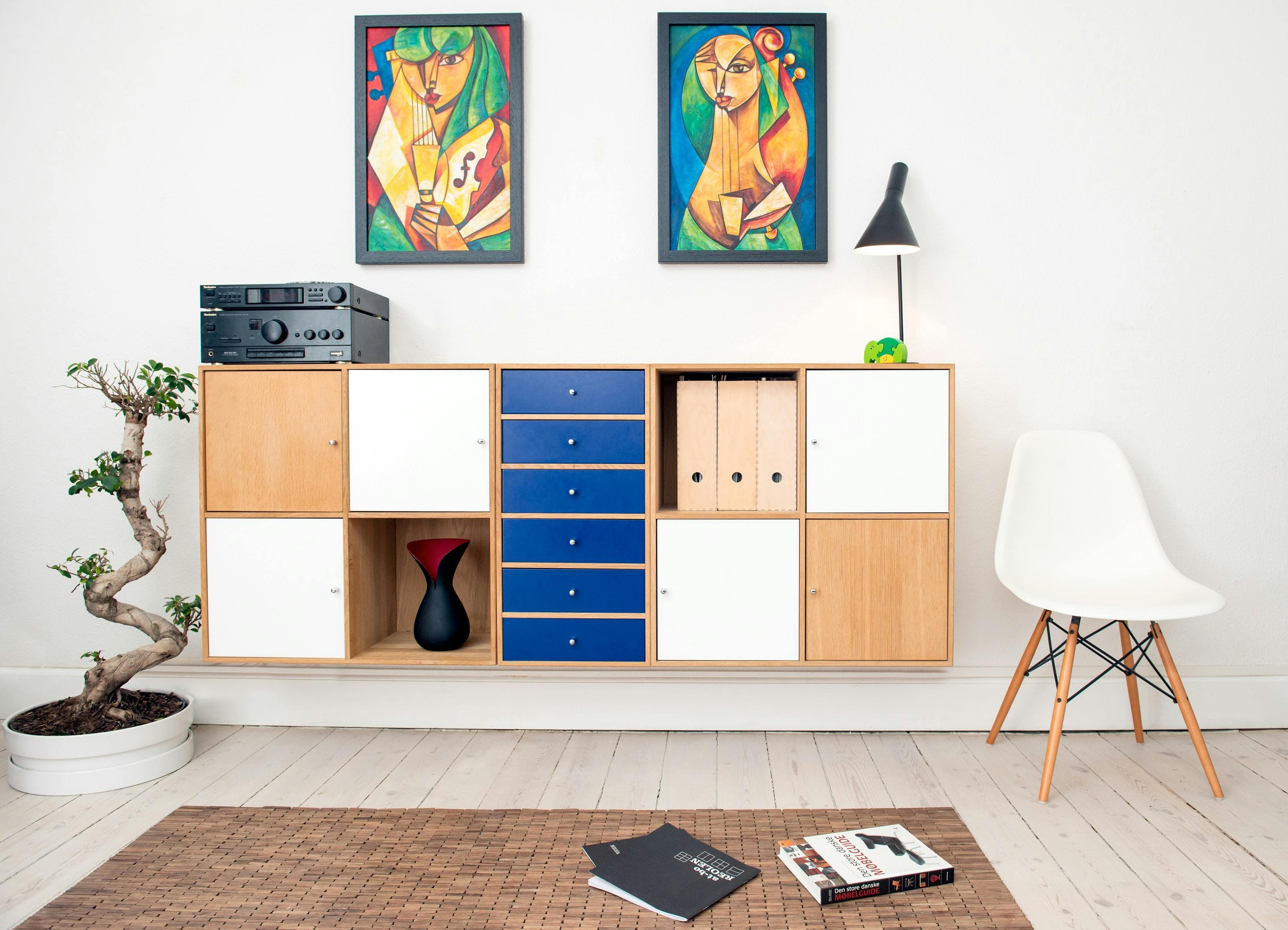 Modern eingerichtetes Wohnzimmer mit einem hängenden Sideboard in Holz, Weiß und Blau, zwei abstrakten Gemälden an der Wand, einem weißen Designerstuhl, einem Bonsai-Baum und dekorativen Elementen wie Büchern und einer schwarzen Vase.