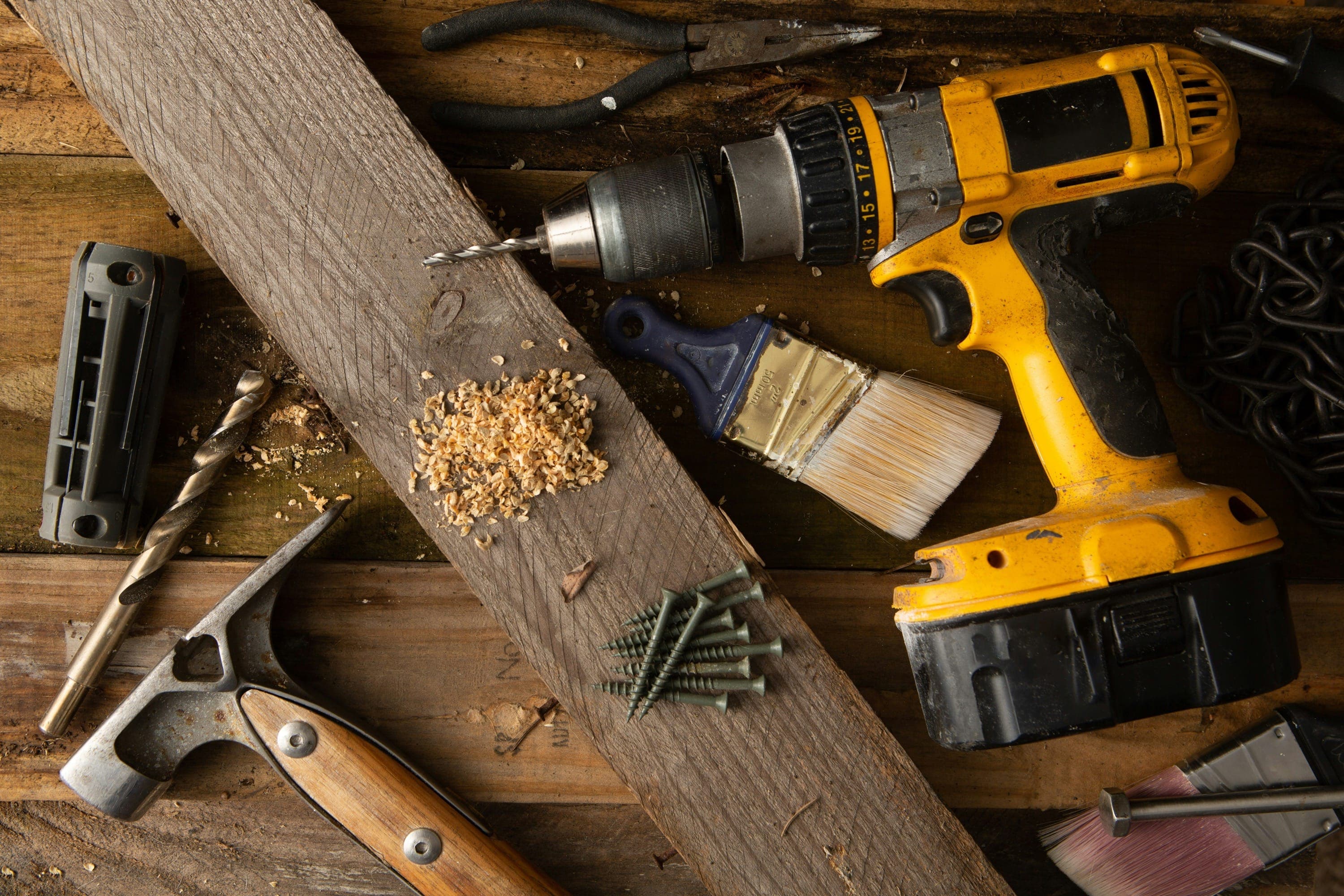 Verschiedene Werkzeuge für Holzarbeiten, darunter eine gelbe Akkubohrmaschine, ein Hammer mit Holzgriff, Schrauben, ein Pinsel, Bohrer und eine Zange auf einer rustikalen Holzoberfläche.