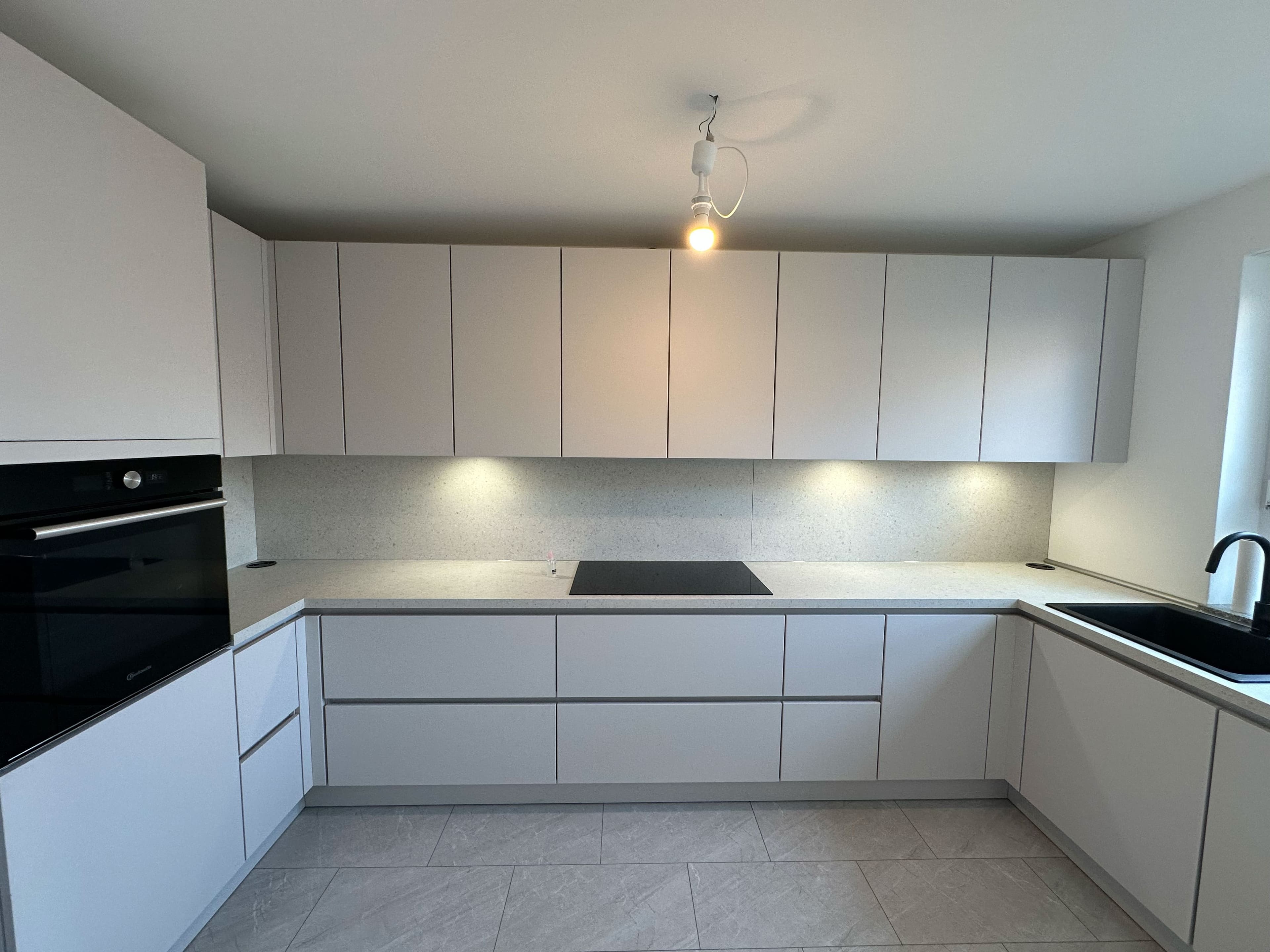 Moderne, vollständig montierte Küche mit griff­losen, lichtgrauen Fronten, integrierter Beleuchtung unter den Oberschränken, schwarzer Spüle und Induktionskochfeld – klare Linien und minimalistisches Design.