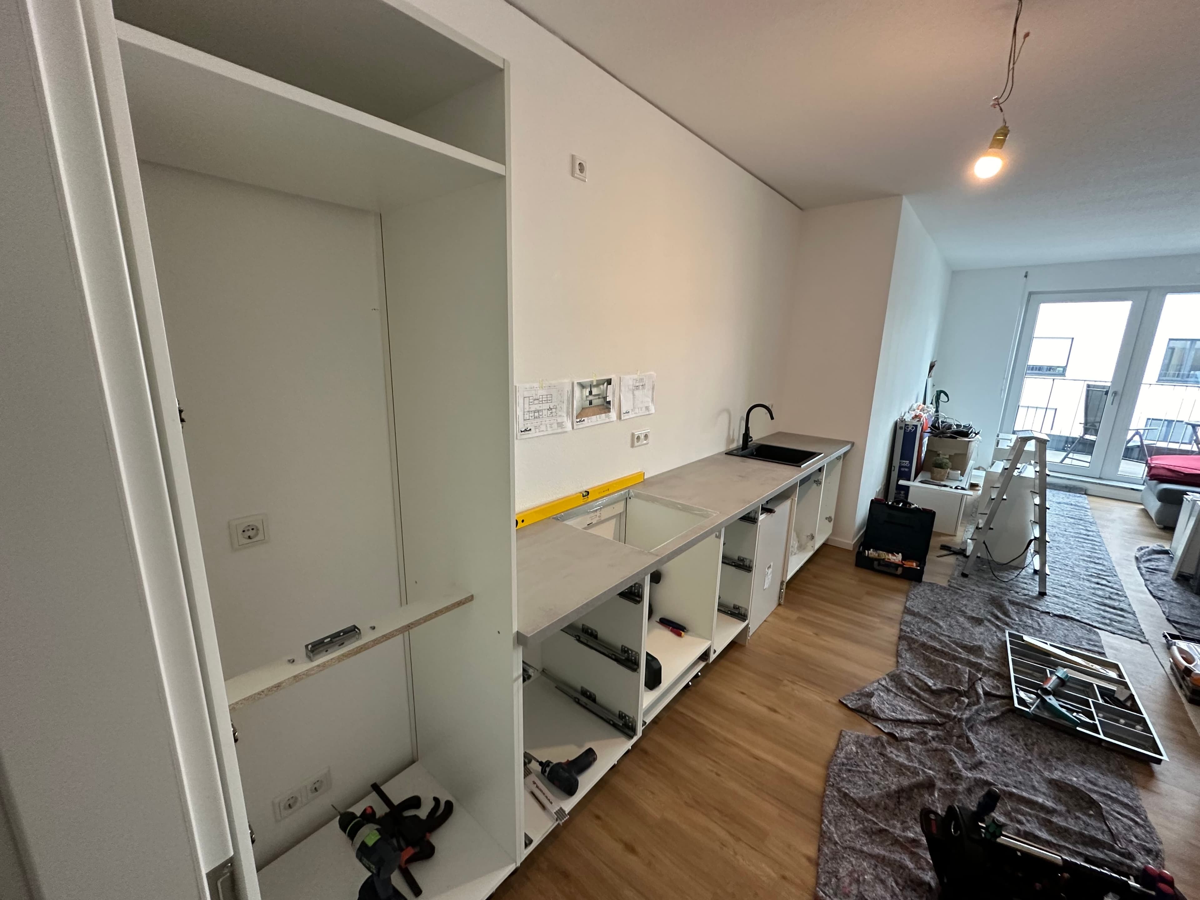 Offene Küchenmontage in einem modernen Wohnraum mit weißen Schränken, Werkzeugen und Abdeckmaterial auf dem Boden – eine Küche im Aufbau mit sichtbaren Anschlüssen und Bauplänen an der Wand.