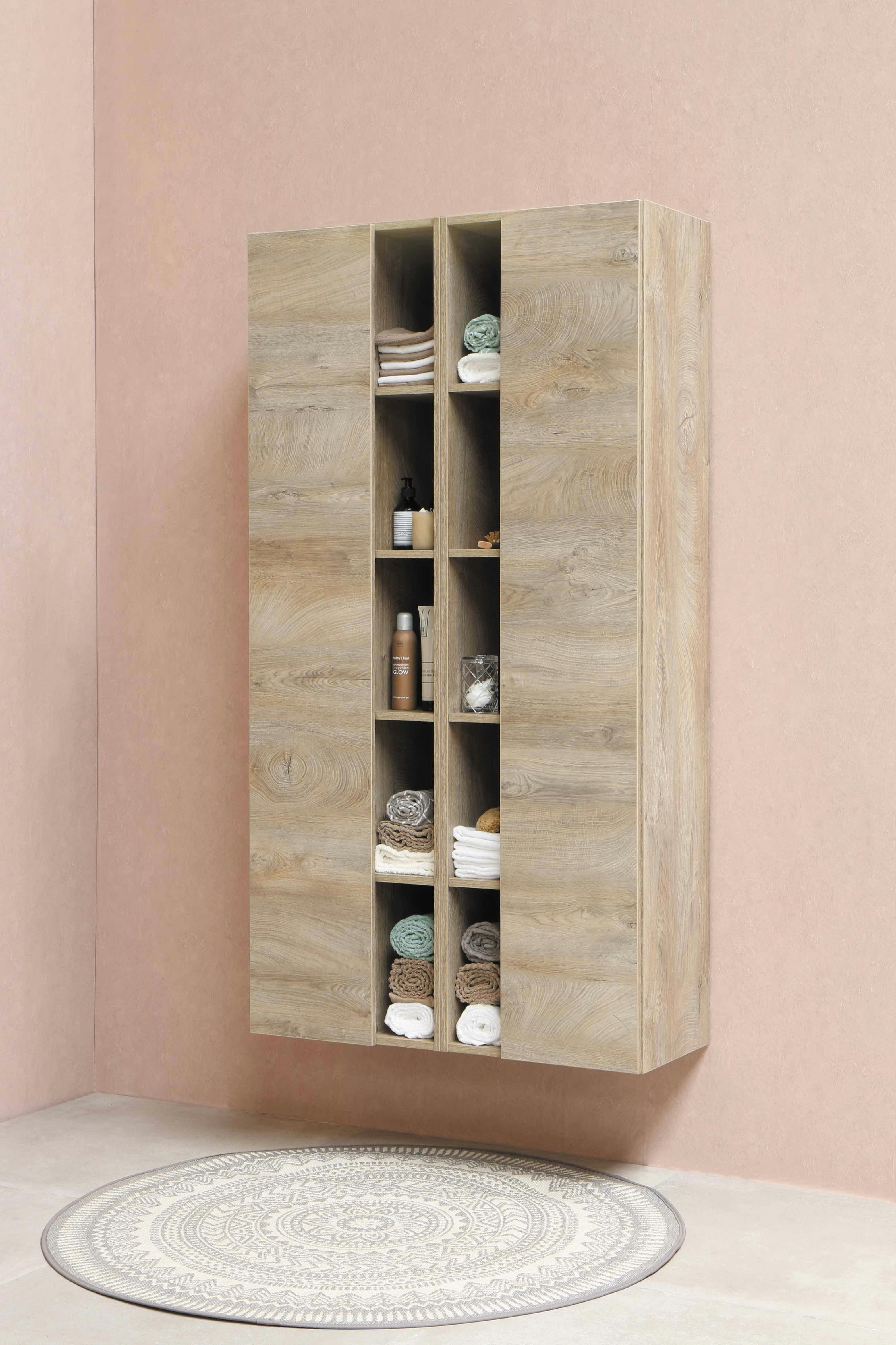 Moderner, wandhängender Badezimmerschrank aus Holz mit offenen Fächern, gefüllt mit Handtüchern, Pflegeprodukten und Dekoration, vor einer rosafarbenen Wand und rundem Teppich mit Ornamentmuster.