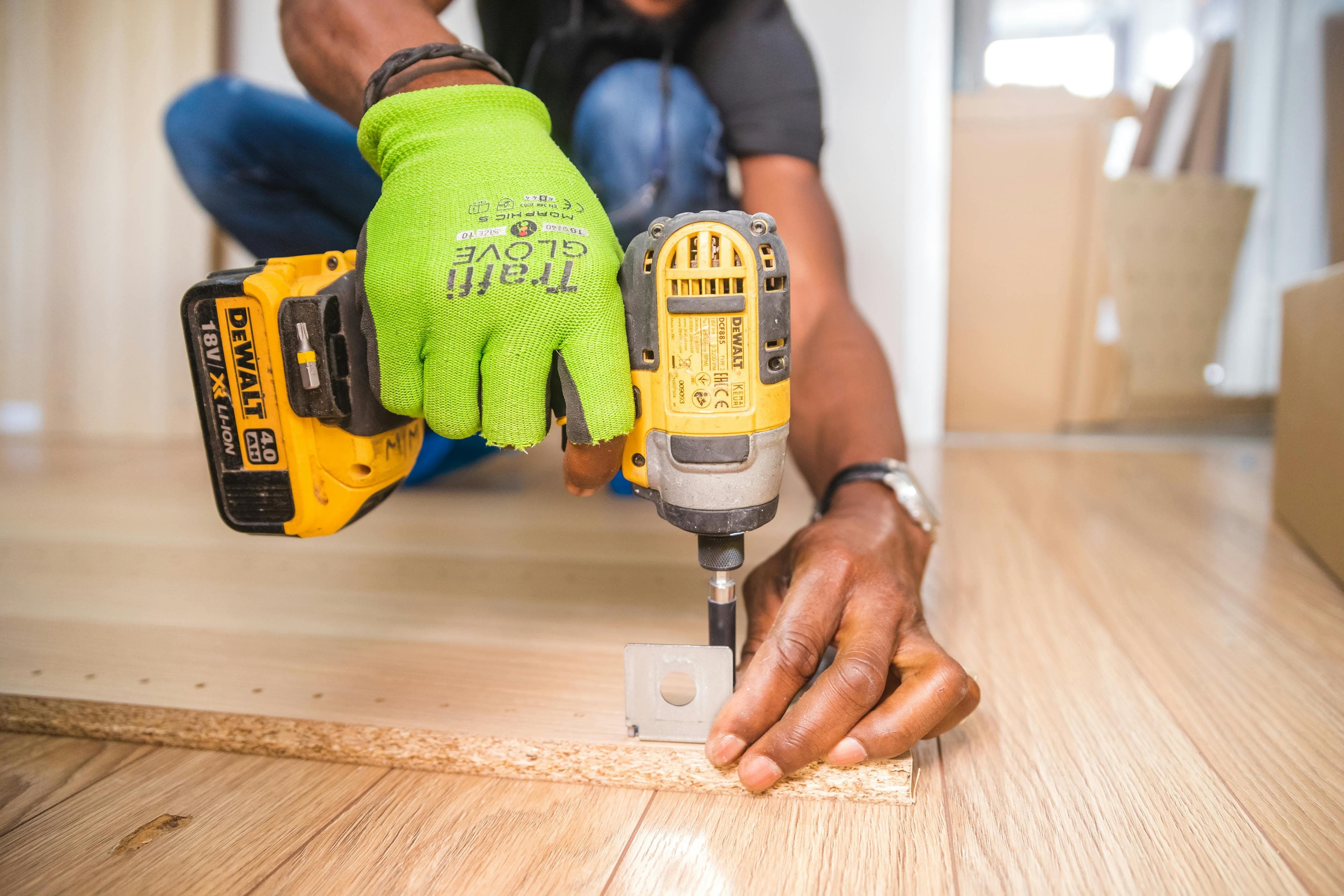Handwerker bei der Möbelmontage, der mit einem gelben Akkuschrauber und grünen Arbeitshandschuhen eine Schraube in eine Spanplatte dreht, um ein Metallteil zu befestigen – auf einem hellen Holzboden in einem Innenraum.